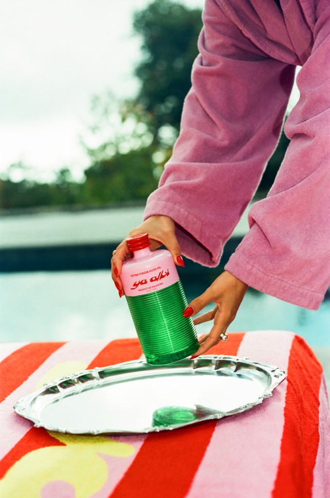 A person in a pink robe is holding a bottle of green liquid labeled 'extra virgin olive oil' above a silver tray placed on a colorful towel by a pool.